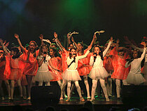 Eine grün beleuchtete Bühne voller winkender Kinder in weißen und flamingofarbenen Tanzkostümen.